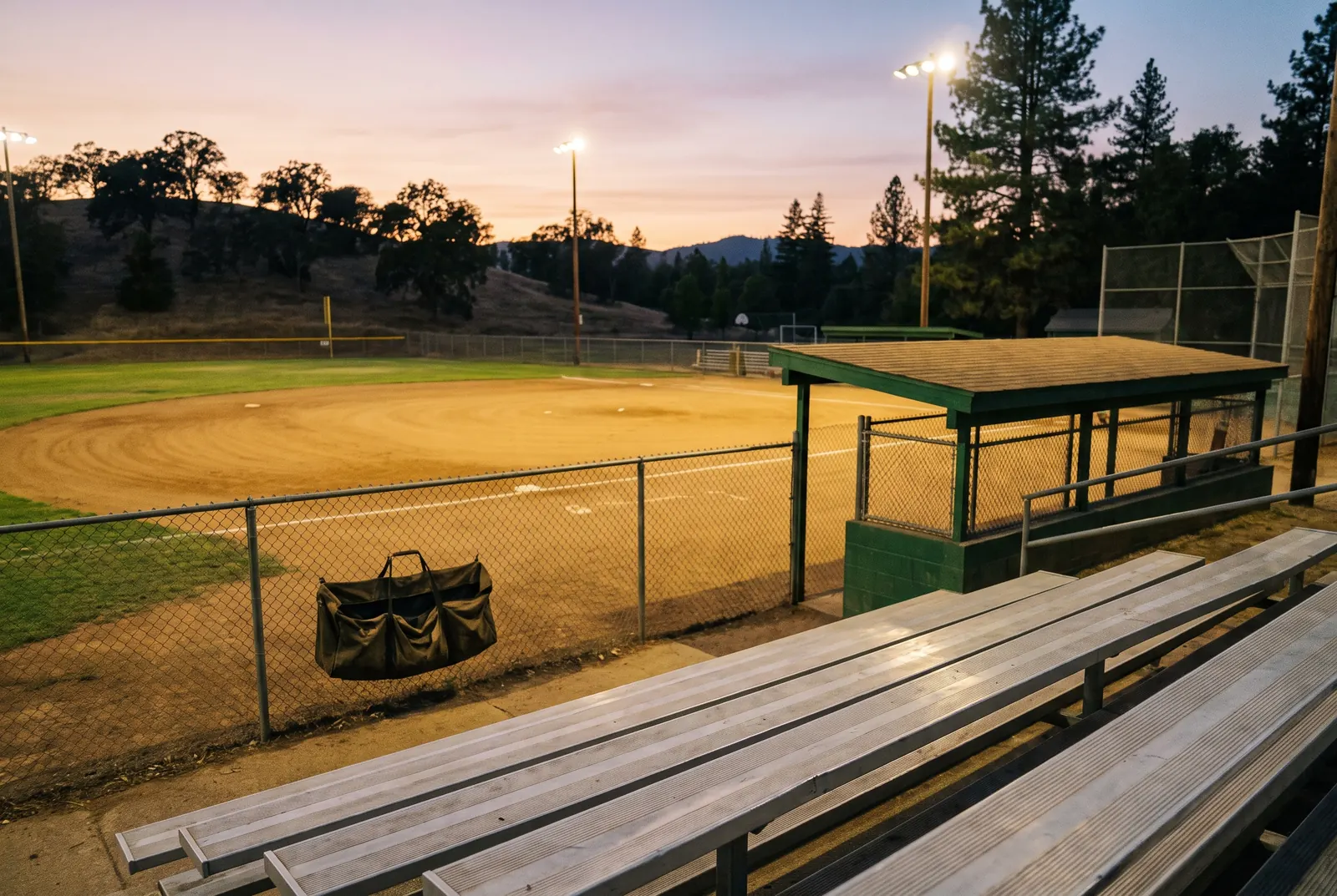 An empty ballfield at dusk seen from the bleachers, the field lights just coming on over the foothills